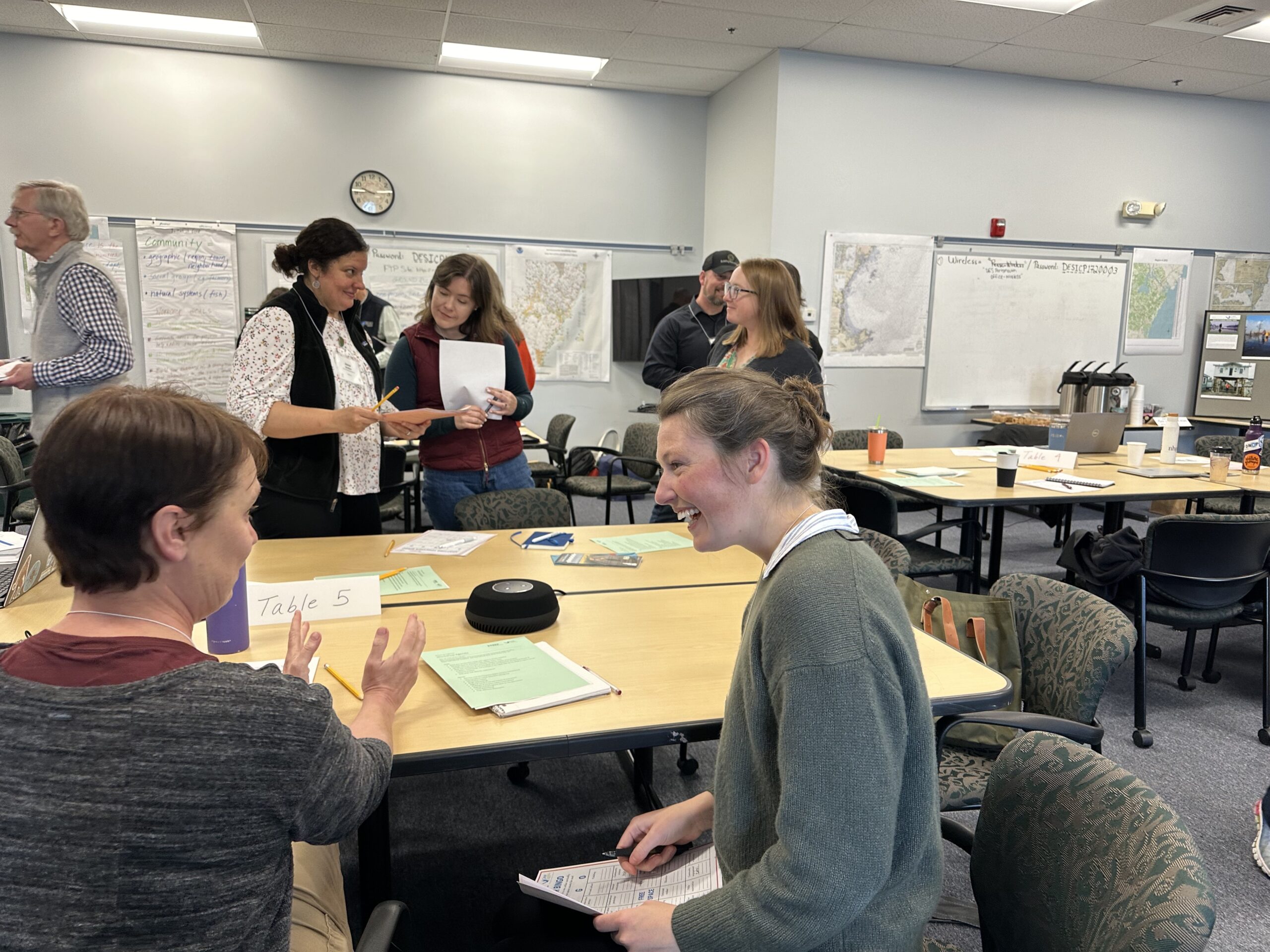 Two people talking in the foreground at a table during the Adapting Together workshop. Several others talk with each other in the background.