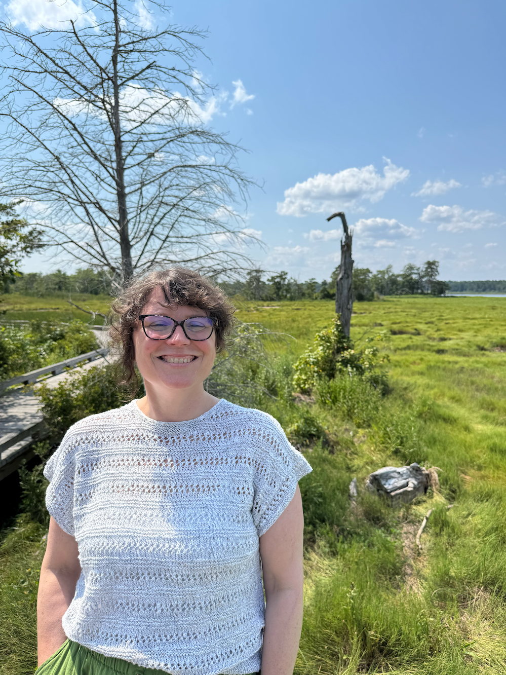 Annie Cox, a white woman with brown hair stands near a salt marsh near Great Bay estuary.