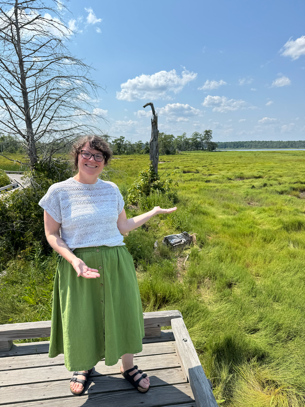 Annie Cox, an adult woman in a green skirt and glasses, smiles and gestures toward Great Bay and verdant salt marshes.