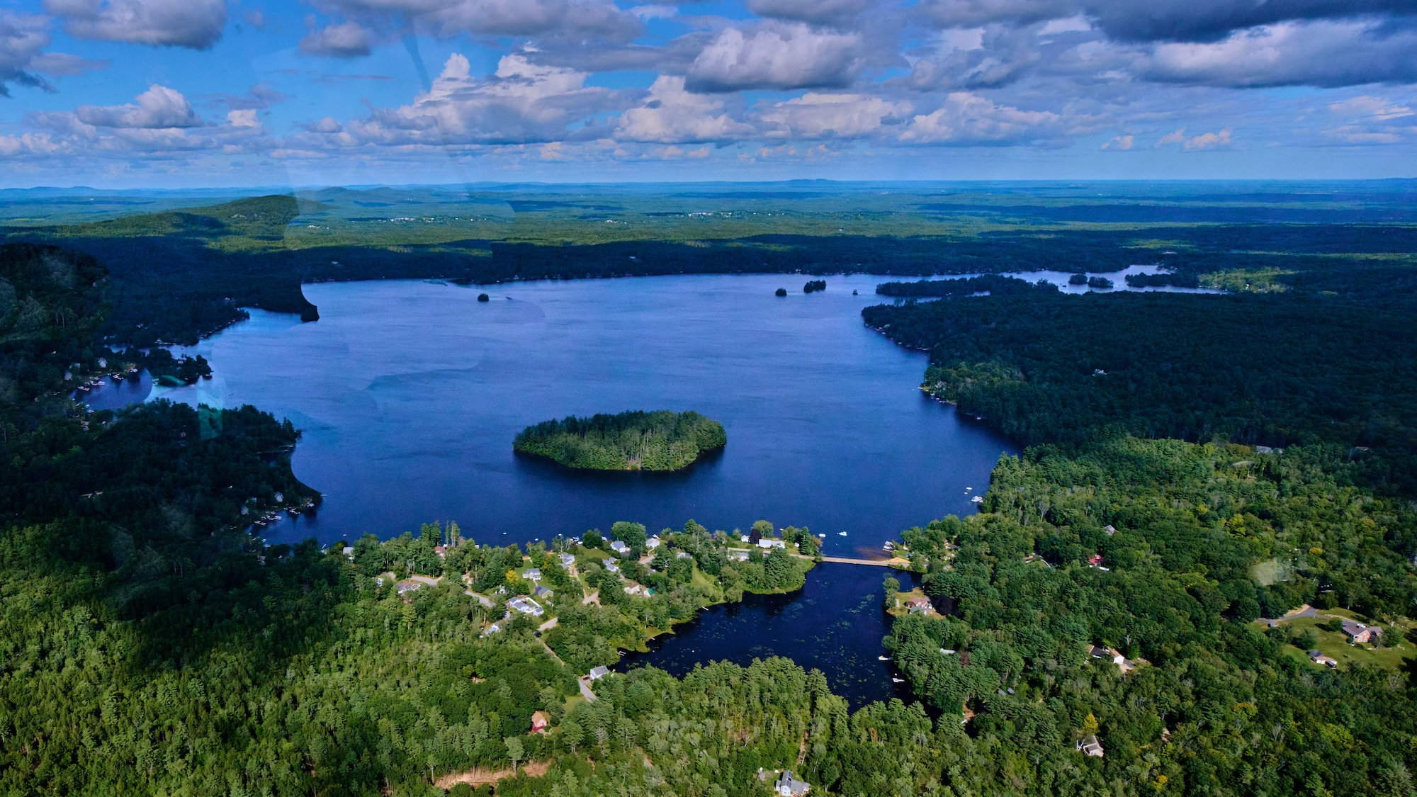 An aerial view of Bow Lake in Strafford, NH. The photo features summer green trees,blue water, and blue sky with puffy clouds. Photo by Robert Bennett.