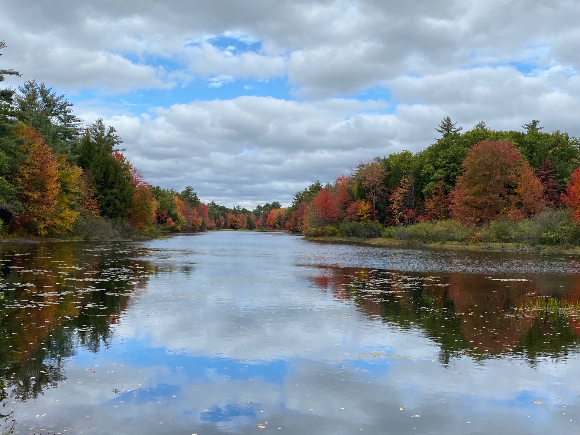 A scenic view of the Exeter River in Fremont, NH. Trees have orange and red fall foliage and blue sky and clouds reflect in the water. Photo by Nancy Murray.
